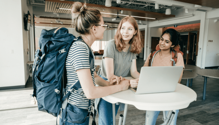 Students with Backpacks Talk Around a Laptop and Small Round Table